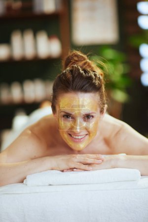 Foto de Tiempo de atención médica. sonriente mujer moderna en el salón de spa con máscara cosmética dorada en la cara puesta en la mesa de masaje. - Imagen libre de derechos