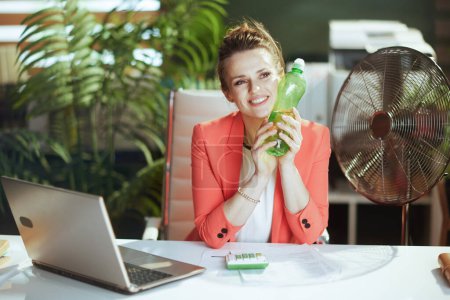 Foto de Lugar de trabajo sostenible. sonriente mujer contable moderna de mediana edad en moderna oficina verde en chaqueta roja con botella de agua, ventilador eléctrico y portátil. - Imagen libre de derechos