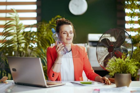 Téléchargez les photos : Un lieu de travail durable. souriant moderne d'âge moyen comptable femme au travail en veste rouge avec ventilateur électrique et ordinateur portable. - en image libre de droit