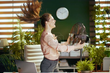 Téléchargez les photos : Un lieu de travail durable. femme d'affaires moderne souriante dans un bureau vert moderne avec ventilateur électrique. - en image libre de droit