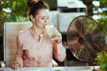 Téléchargez les photos : Un lieu de travail durable. femme d'âge moyen moderne employée au travail avec ventilateur électrique, tasse d'eau et citron. - en image libre de droit