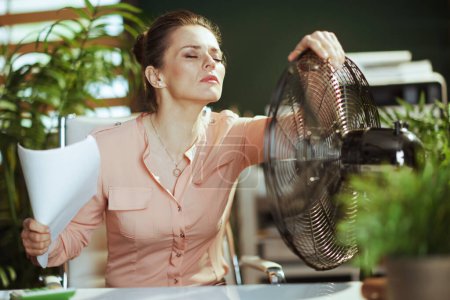 Sustainable workplace. modern accountant woman in modern green office with documents and electric fan suffering from summer heat.