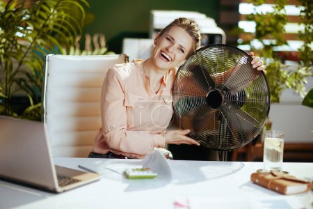 Foto de Lugar de trabajo sostenible. contenta mujer contable moderna de mediana edad en la oficina verde moderna con ventilador eléctrico y portátil. - Imagen libre de derechos