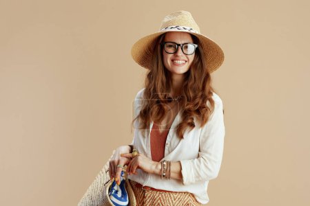 Téléchargez les photos : Vacances à la plage. sourire élégante femme de 40 ans en chemisier blanc et short isolé sur beige dans des lunettes avec sac de paille et chapeau d'été. - en image libre de droit