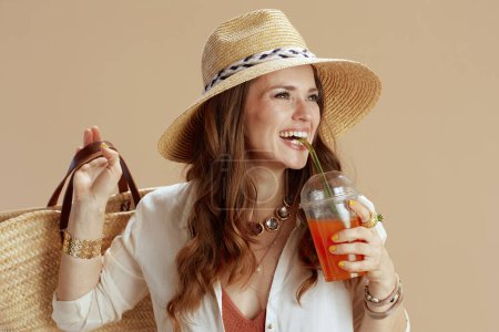 Photo for Beach vacation. smiling elegant 40 years old woman in white blouse and shorts isolated on beige background with straw bag, carrot juice and summer hat. - Royalty Free Image