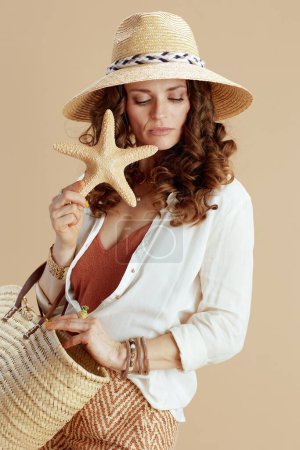 Photo for Beach vacation. pensive elegant woman in white blouse and shorts isolated on beige with sea star, straw bag and summer hat. - Royalty Free Image
