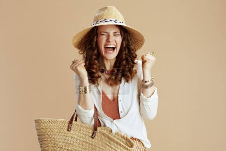 Téléchargez les photos : Vacances à la plage. femme moderne et souriante d'âge moyen en chemisier blanc et short sur fond beige avec sac en paille et chapeau en paille avec bras levés se réjouissant. - en image libre de droit
