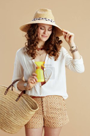 Foto de Vacaciones en playa. sonriente hembra de moda en blusa blanca y pantalones cortos aislados en beige con bolsa de paja, spf y sombrero de paja. - Imagen libre de derechos