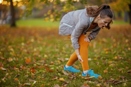 Photo for Hello autumn. unhappy modern middle aged woman in fitness clothes in the park with ankle pain. - Royalty Free Image