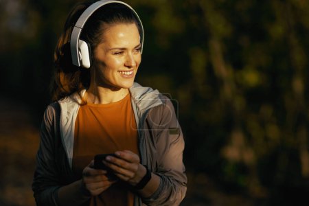 Photo for Hello autumn. happy stylish middle aged woman in fitness clothes in the park listening to the music with headphones. - Royalty Free Image
