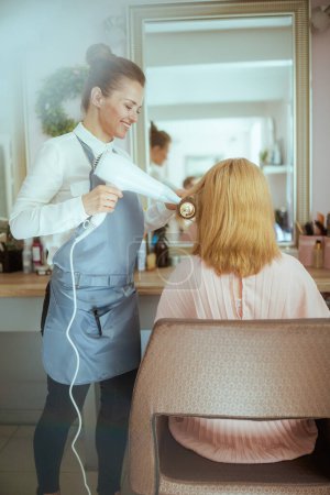 middle aged woman hairdresser in modern beauty salon with hairbrush and client blowout hair with hair dryer.