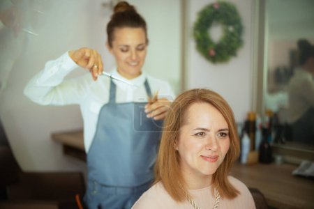 Téléchargez les photos : Femme de 40 ans coiffeuse dans un salon de beauté moderne avec ciseaux et client coupe les cheveux. - en image libre de droit