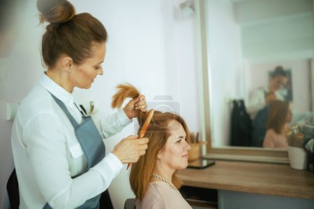 Téléchargez les photos : 40 ans employé de salon de coiffure dans un studio de coiffure moderne avec brosse à cheveux et client. - en image libre de droit