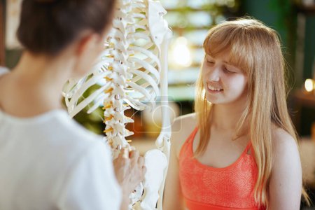Healthcare time. happy female medical massage therapist in massage cabinet with teenage client and skeleton.