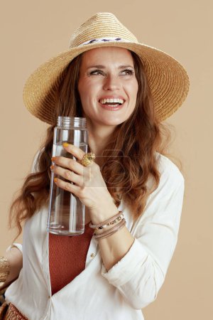 Téléchargez les photos : Vacances à la plage. heureuse femme élégante en chemisier blanc et short isolé sur fond beige avec bouteille d'eau et chapeau d'été. - en image libre de droit