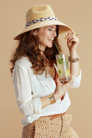 Téléchargez les photos : Vacances à la plage. heureuse femme d'âge moyen moderne en chemisier blanc et short isolé sur fond beige avec cocktail et chapeau de paille. - en image libre de droit