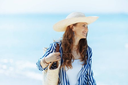 Foto de Feliz mujer moderna de 40 años en la costa del océano con bolsa de paja y sombrero de paja. - Imagen libre de derechos