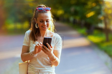 Foto de Hora de verano. sonriente mujer de mediana edad de moda en camisa blanca con bolso de mano usando teléfono inteligente y caminando en el parque de la ciudad. - Imagen libre de derechos