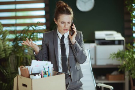 Un nuevo trabajo. acentuó la trabajadora moderna de mediana edad en la oficina verde moderna en traje de negocios gris con pertenencias personales en caja de cartón hablando en un teléfono inteligente.