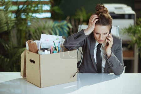 Photo for New job. stressed modern female worker in modern green office in grey business suit with personal belongings in cardboard box talking on a smartphone. - Royalty Free Image