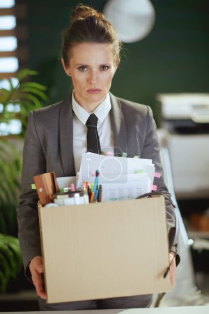 Un nuevo trabajo. infeliz moderno 40 años de edad, trabajadora en moderna oficina verde en traje de negocios gris con pertenencias personales en caja de cartón.