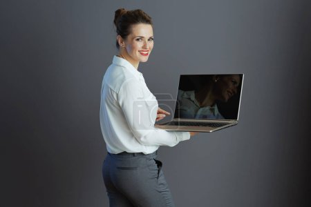Téléchargez les photos : Femme d'affaires élégante et souriante en chemisier blanc avec ordinateur portable isolé sur fond gris. - en image libre de droit