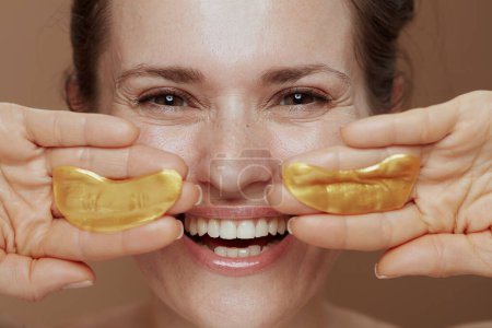 Téléchargez les photos : Femme d'âge moyen moderne souriante avec des patchs pour les yeux sur fond beige. - en image libre de droit