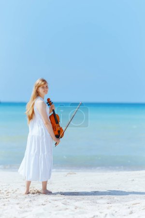 Retrato de larga duración de una adolescente moderna sonriente vestida de blanco en la costa del océano con violín.