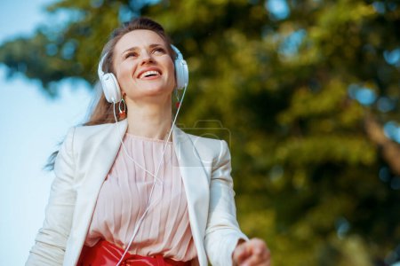 smiling modern woman in pink dress and white jacket in the city with headphones.