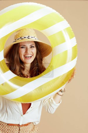 Téléchargez les photos : Vacances à la plage. femme élégante souriante en chemisier blanc et short isolé sur fond beige avec anneau gonflable et chapeau de paille. - en image libre de droit