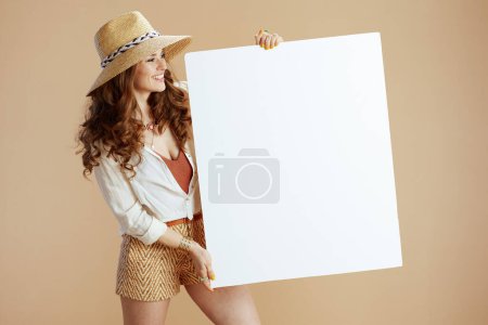 Photo for Beach vacation. smiling stylish 40 years old woman in white blouse and shorts on beige background with straw hat showing blank board. - Royalty Free Image