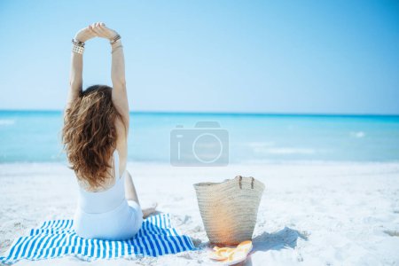 Photo for Seen from behind elegant woman on the ocean coast with straw bag and striped towel. - Royalty Free Image