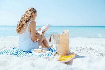 Foto de Mujer de mediana edad elegante relajado en la playa con bolsa de paja, libro y toalla a rayas. - Imagen libre de derechos