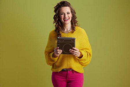 glücklich gesunde Frau mittleren Alters isoliert auf grünem Hintergrund in gelbem Pullover mit Tablet-PC.
