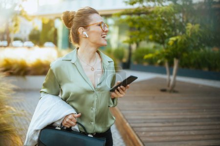 Photo for Happy modern 40 years old business woman near office building in green blouse and eyeglasses with briefcase and wireless headphones using smartphone and walking. - Royalty Free Image
