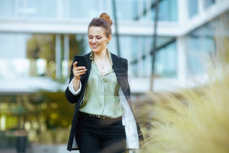 lächelnde moderne Geschäftsfrau mittleren Alters im Geschäftsviertel in schwarzer Jacke mit Smartphone zu Fuß.
