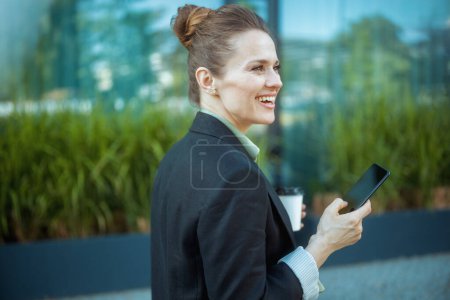 heureuse femme d'affaires moderne près du centre d'affaires en veste noire avec smartphone et tasse de café.