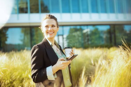 Photo for Smiling modern 40 years old woman employee near business center in black jacket with cup of coffee using smartphone. - Royalty Free Image