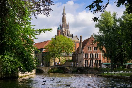 Foto de Bruges, Belgium-July 4, 2022: A canal in Bruges with a church in the distance, summer, park - Imagen libre de derechos