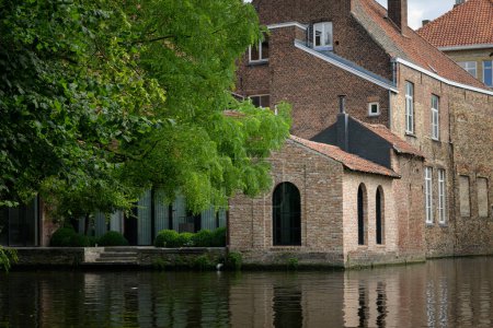 Foto de Bruges, Belgium-July 4, 2022: An old house at a canal with a bridge, daylight, trees - Imagen libre de derechos