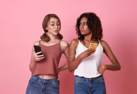 Foto de Joven dos mujeres usando el teléfono móvil mientras que la mujer morena mira el teléfono inteligente de su amigo en el fondo rosa. privacidad secreta en las redes sociales. - Imagen libre de derechos