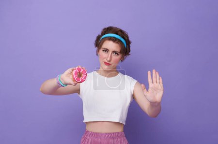 Young sad woman hold eat burger show hand stop gesture say no junk food isolated on purple background. Proper nutrition healthy lifestyle fast food unhealthy choice concept