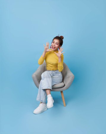 Foto de Emocionado asiático adolescente chica sentado en silla y abierto boca gritando anunciar aislado en azul fondo Wow y sorprendido concepto - Imagen libre de derechos