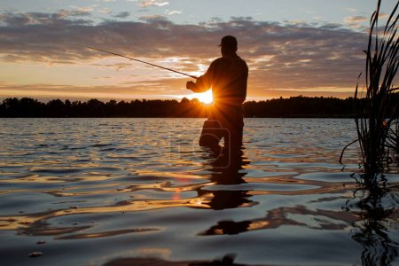 Foto de Pescador con un wader pecho de pie en el lago de pesca al atardecer con defecto de lente llamarada - Imagen libre de derechos