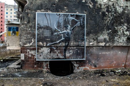 Photo for "Girl gymnast" Mural by Banksy in Irpin, on a ruined building, just above the shell, another little gymnast is painted - this time with a ribbon. Looks like that hole is a ball the girl dances on. - Royalty Free Image