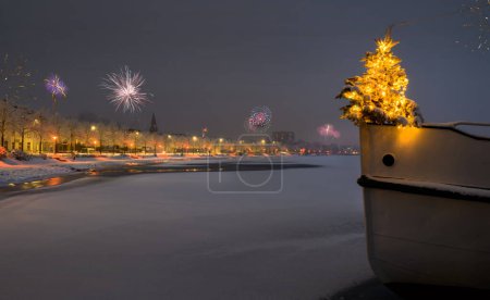 Der Weihnachtsbaum steht am Bugrand eines Bootes und im Hintergrund Feuerwürmer. Silvesterzeit auf der Schlei. Feuerwerk an Neujahr. Winterpromenade in Schleswig mit Schnee bedeckt. 
