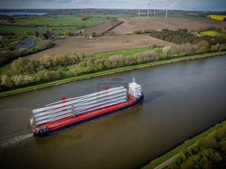 Ship on Kiel Canal loaded with wind turbine blades. Shipping of wind turbine components on cargo ship via the Kiel Canal. Along canal's banks, renewable energy solutions such as solar and wind park.