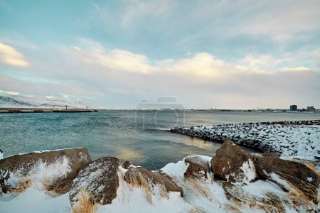 Winter landscape with snow-covered rocks and a calm ocean under a cloudy sky. Location: Reykjavik Iceland.