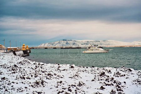 Winterlandschaft mit schneebedeckter Küste und Boot auf kaltem, blauem Wasser mit Bergen im Hintergrund. Standort: Reykjavik Island.
