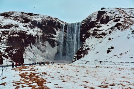 Tourists at a majestic icy waterfall with snow-covered cliffs and a partly cloudy sky. Location: Skogafoss Waterfall Iceland.